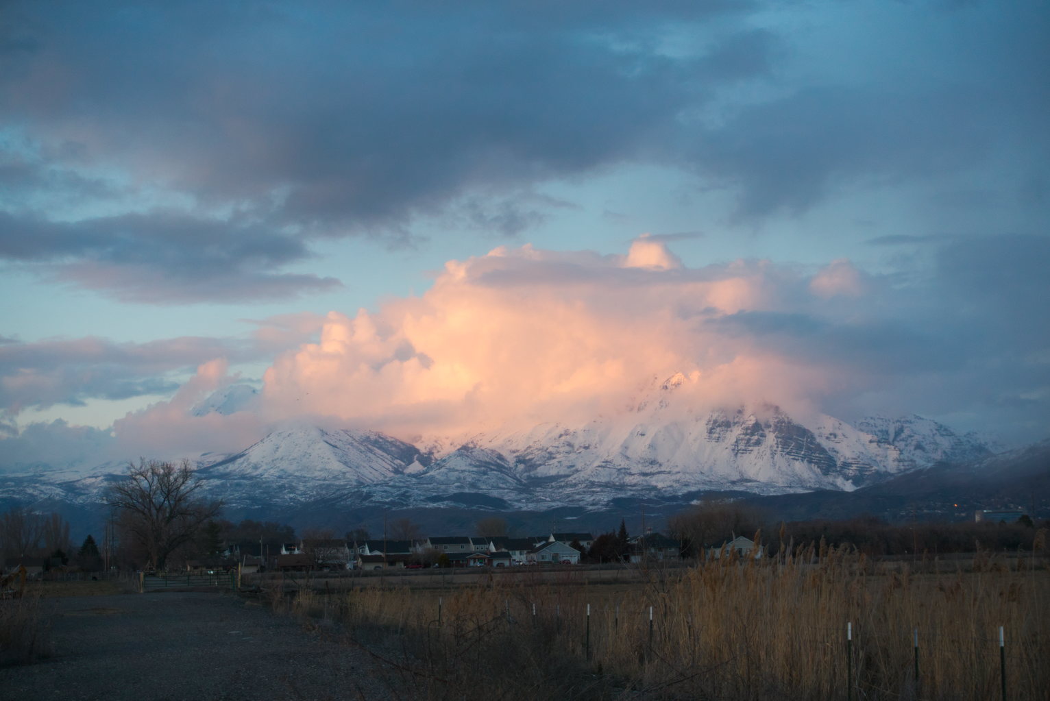 A gravel road, a field, some houses, and above them the snowy mountains engulfed in peachy clouds