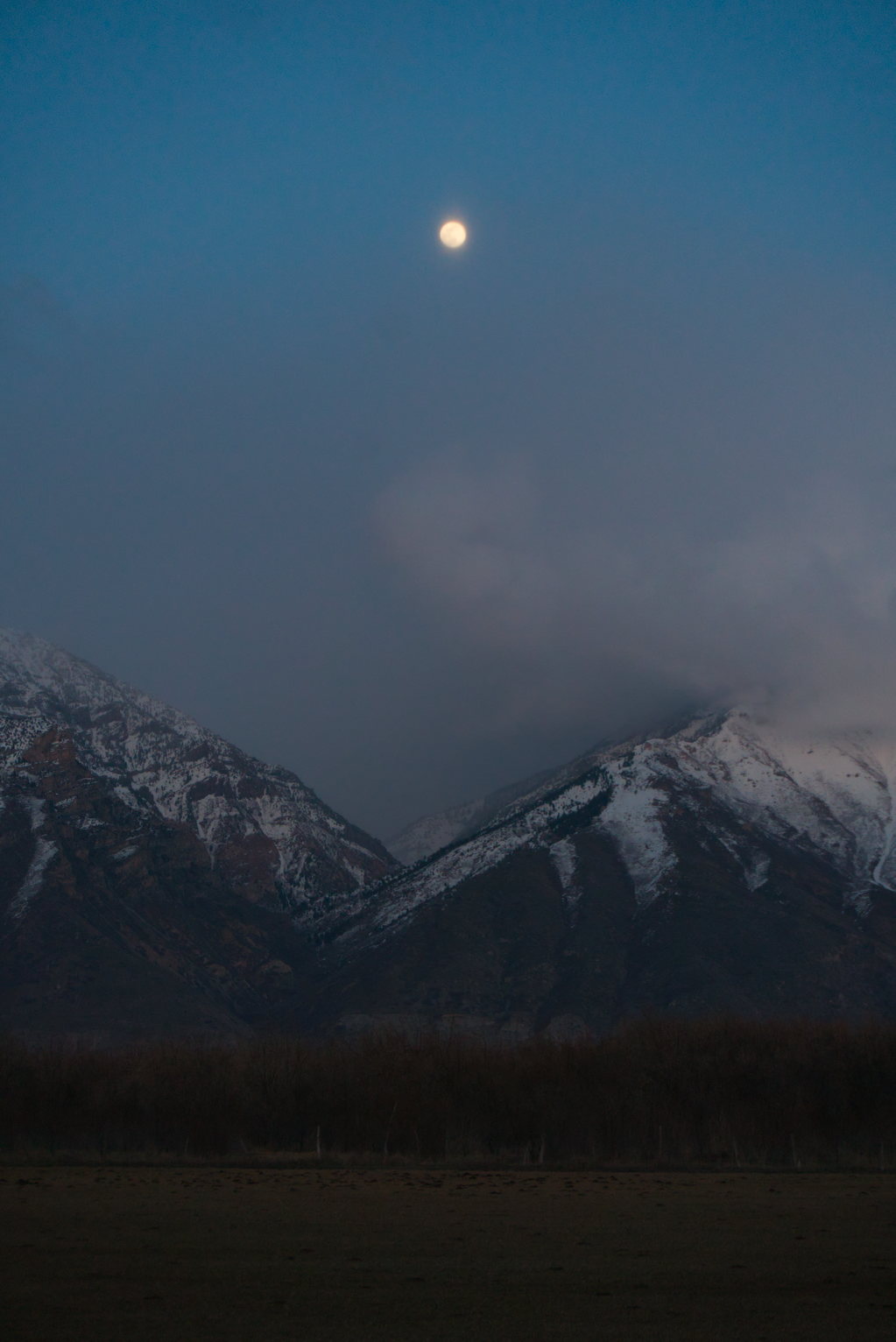 The nearly full moon above snowy mountains at dusk misty with clouds
