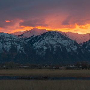 Snowy mountains with orange glowing clouds above, their reflection in some water below, inbetween dry winter grasses