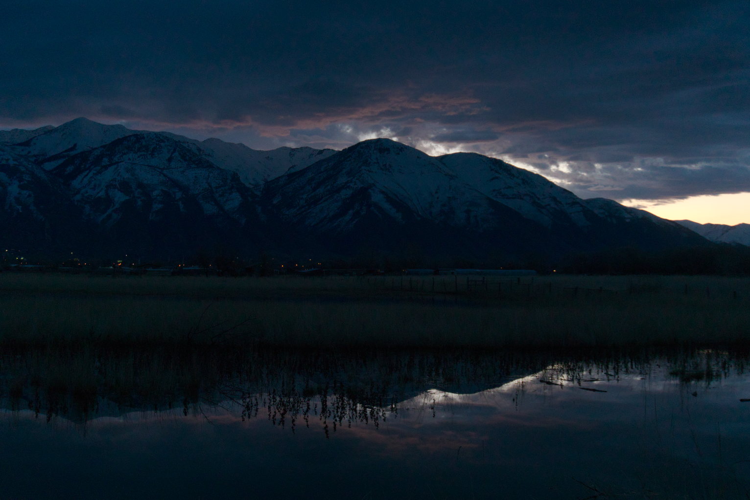 Snowy mountains in the dark, the beginnings of a sunrise visible in the clouds, some reflection in water below