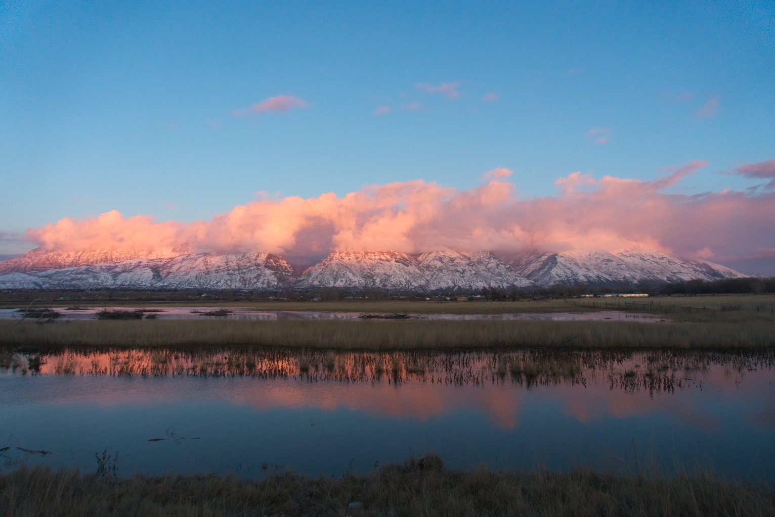 A wide view of the mountains, their tops covered in pinky clouds, and the clouds reflection in teh water below from the wetlands