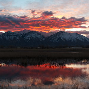 Clouds glowing like embers above snowy mountains at sunrise, reflected in some wetland water in foreground