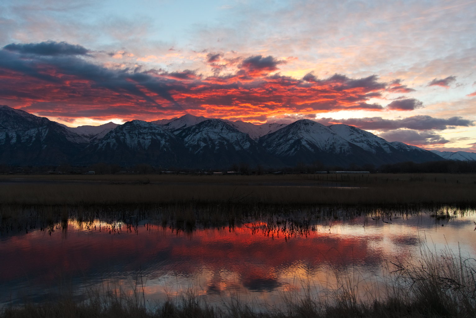 Clouds glowing like embers above snowy mountains at sunrise, reflected in some wetland water in foreground