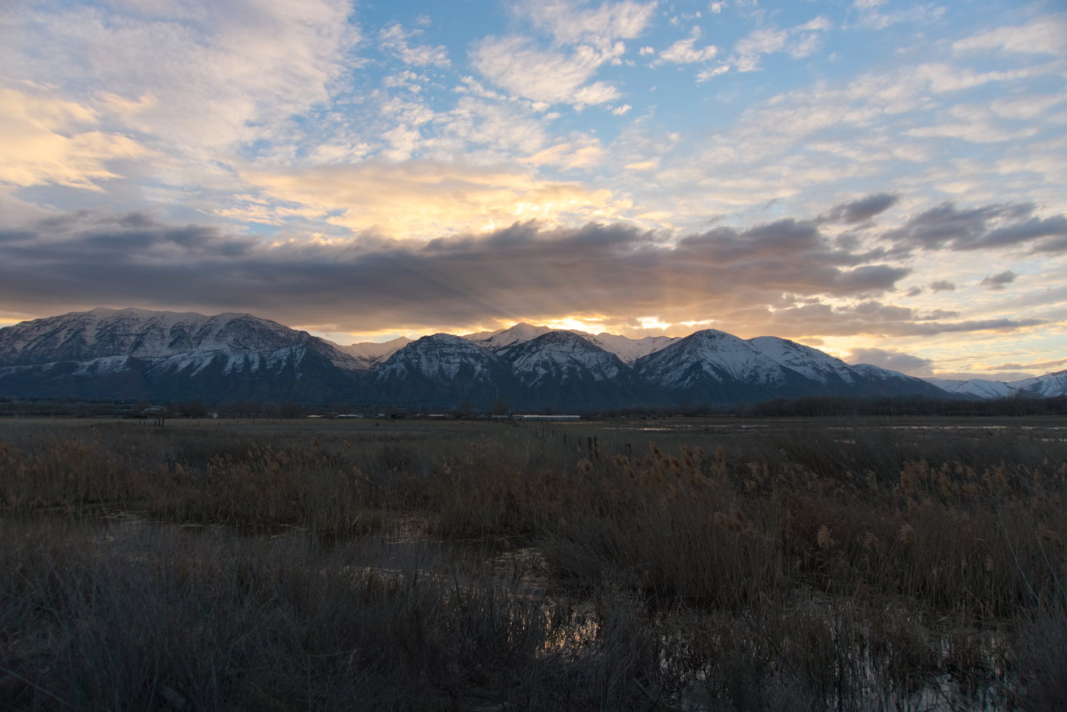 Sunrays burst out from behind the snowy mountains at sunrise, wetlands in the foreground