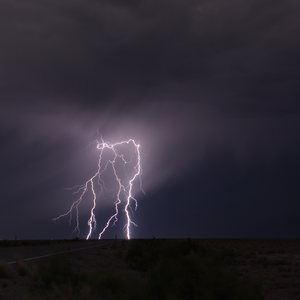Dramatic lightning bolts illuminate dark storm clouds over a rural landscape with a road in the foreground and distant mountains silhouetted against the stormy sky.