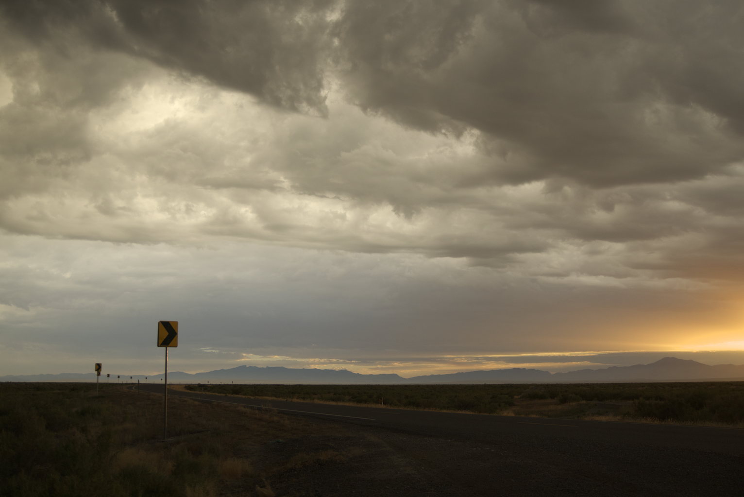 A rural road stretches toward distant mountains under towering storm clouds with a yellow road sign in the foreground and dramatic sky overhead.