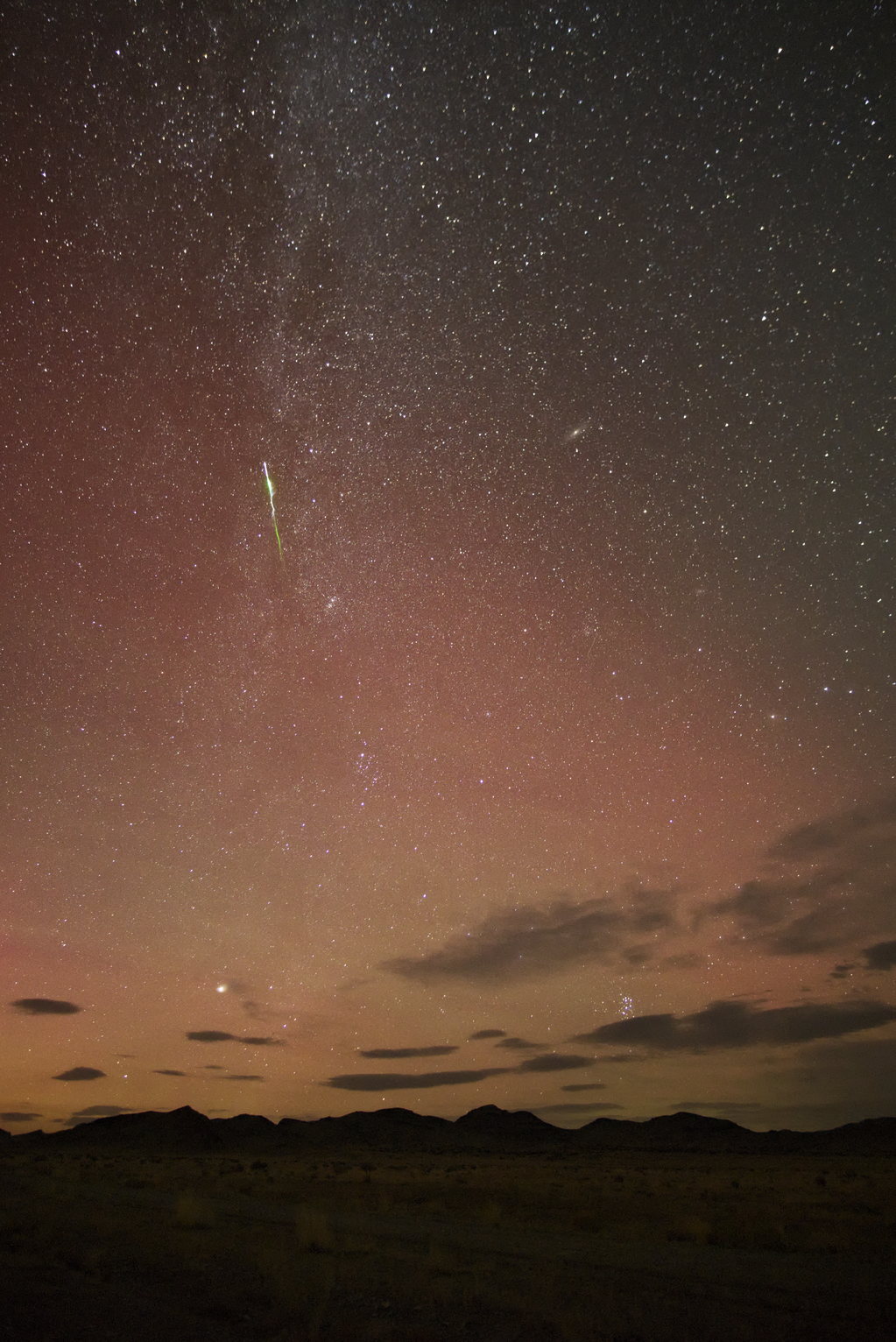 A bright Perseid meteor streaks across the star-filled sky, with the Milky Way visible in the background above a dark landscape silhouette.