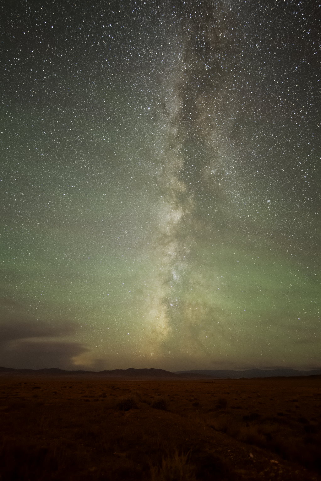 The Milky Way galaxy core glows prominently in a star-dense night sky above rolling hills silhouetted against the starlight. The landscape glows red in aurora light. The sky is green with airglow.