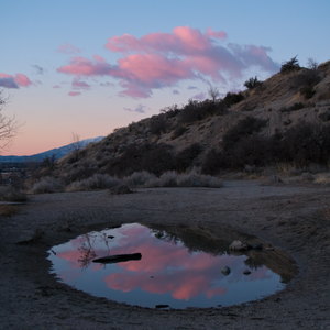 Pink clouds reflected in a pool of water in the dirt on a wintry, brown mountainside