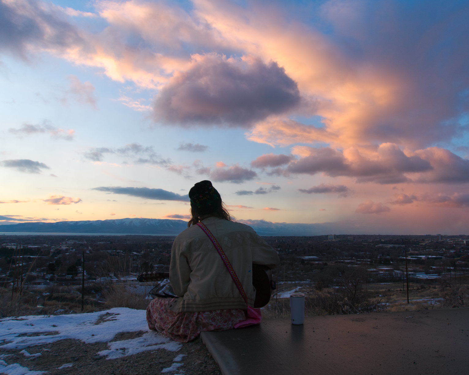 A guitar player sits on the mountainside looking out on the peachy clouds over town.