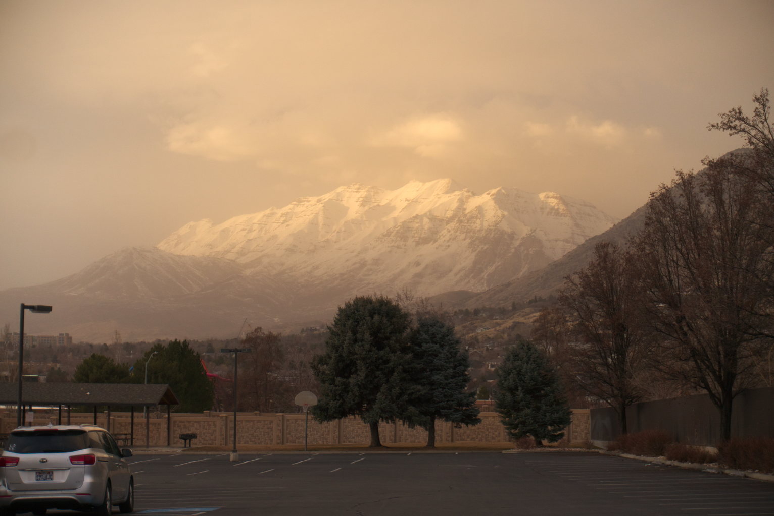 Beyond a mostly empty parking lot and some pines, town and the high snowy mountains hazy in dusty air, catching warm light