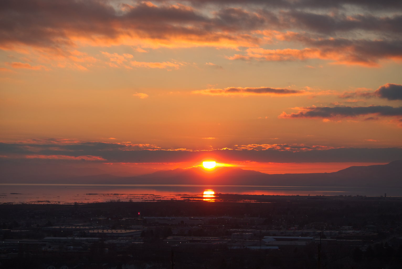 The sun halfway sets below the mountains to the west on the other side of the lake, below part of town, and above the orange lit clouds