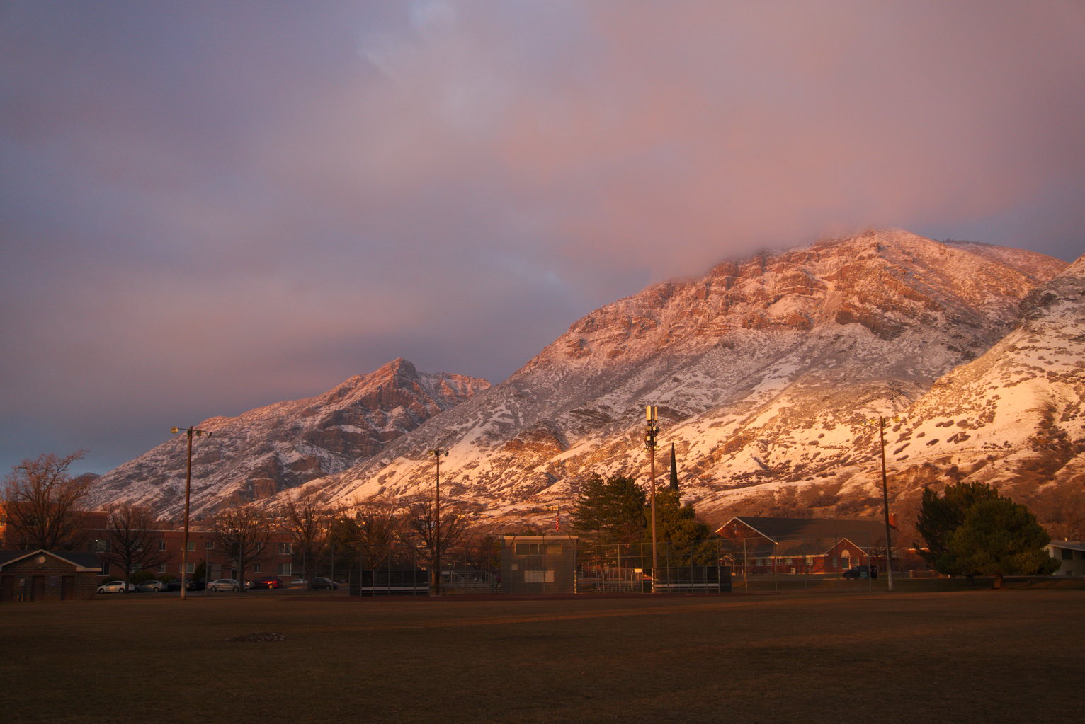 The snowy mountains catching bands of sunset light, viewed from a baseball field, some buildings behind it, and above the mountains some glowy misty looking clouds