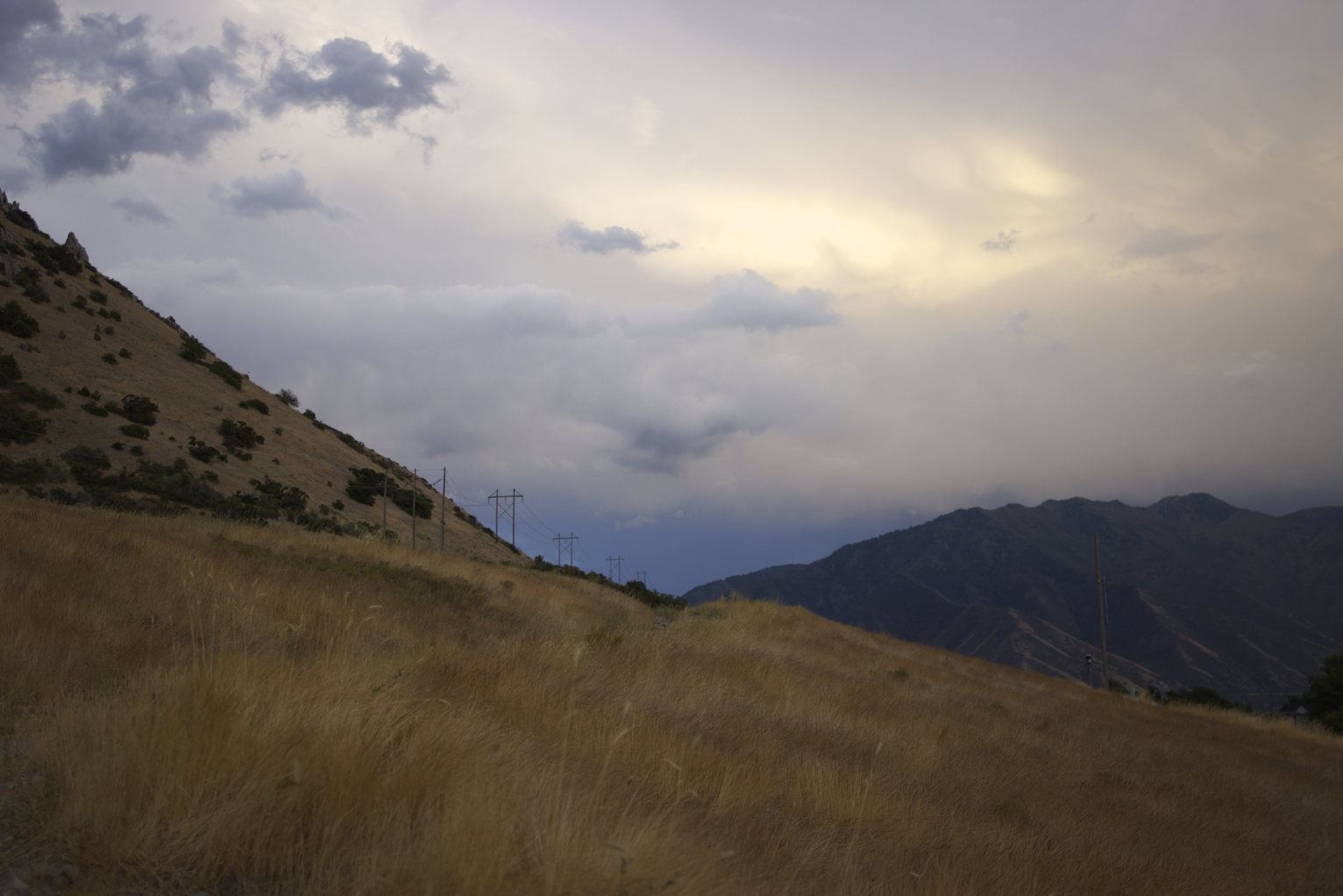 Dry orange-yellow grass on the mountainside, greener mountains beyond, cloudy skues with a glowing spot.