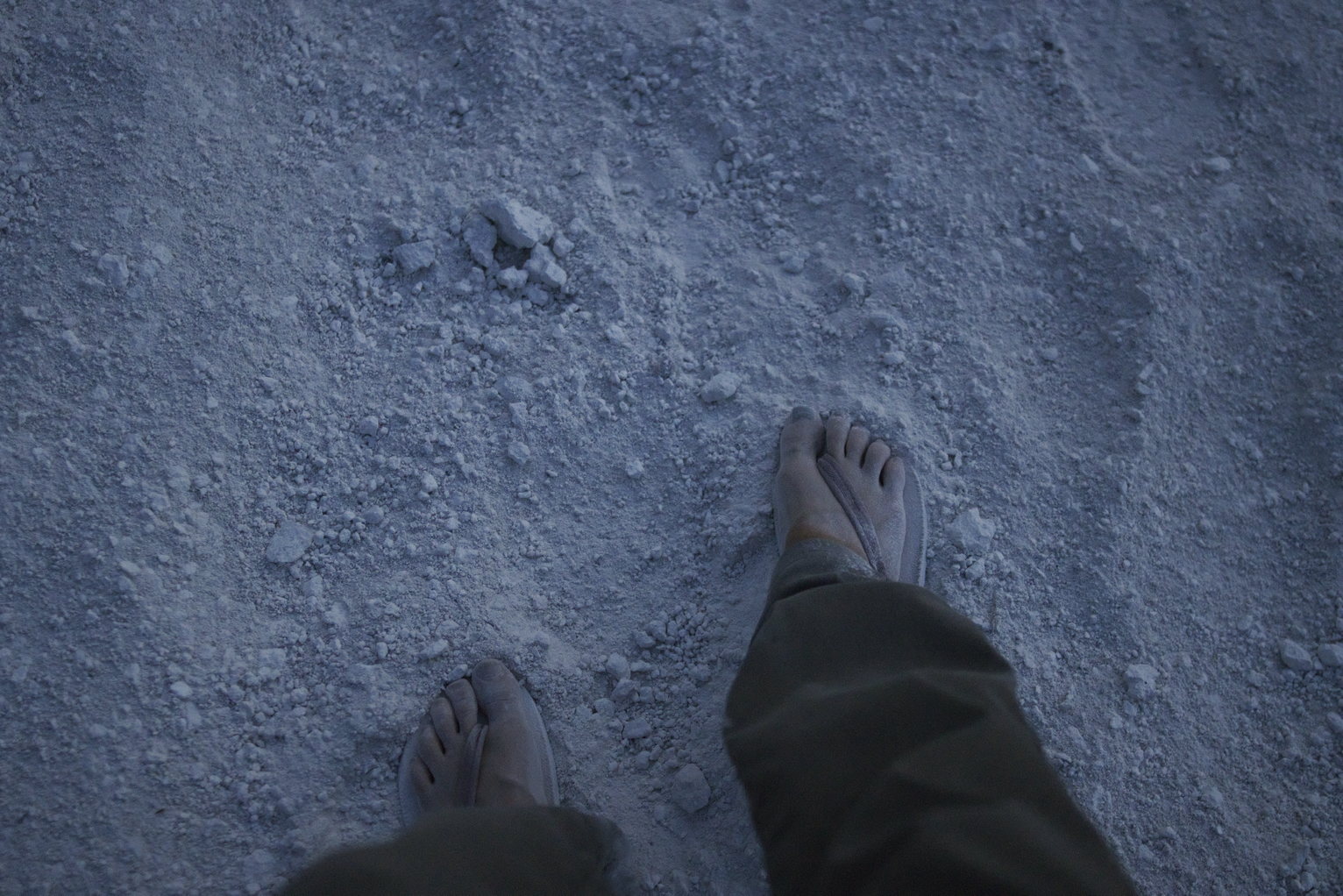 Sandled feet viewed from above on white, pebbly dirt.