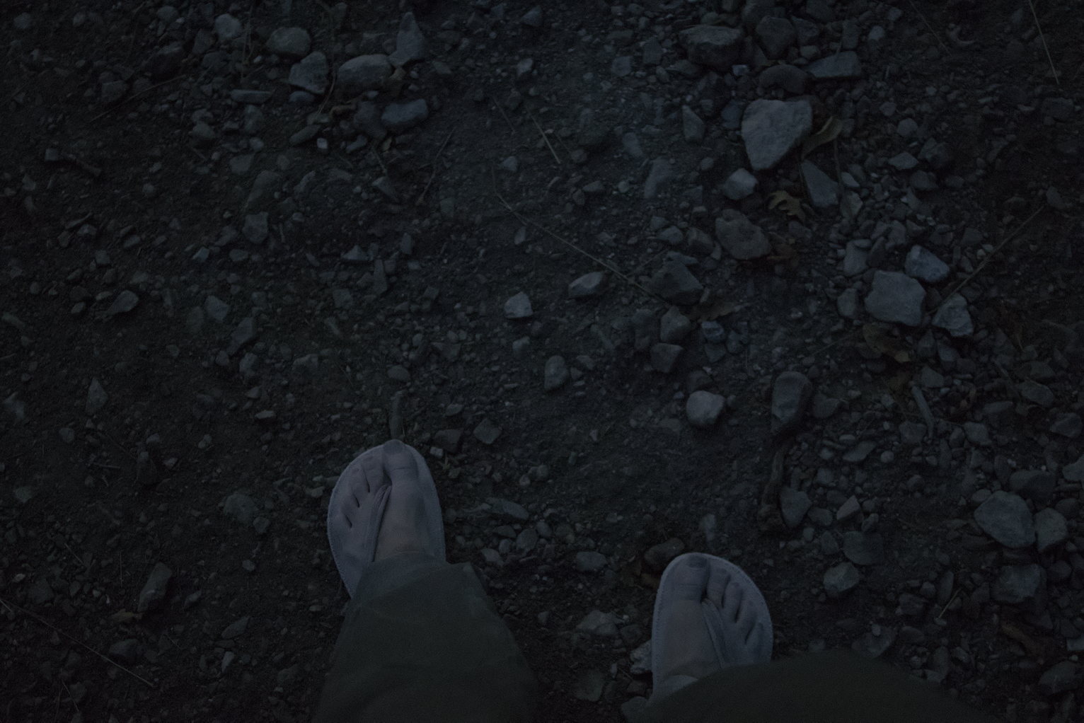 Sandled feet viewed from above all white and powdery as if they'd been dipped in flour and looking ghostly on rocky dirt in twilight.