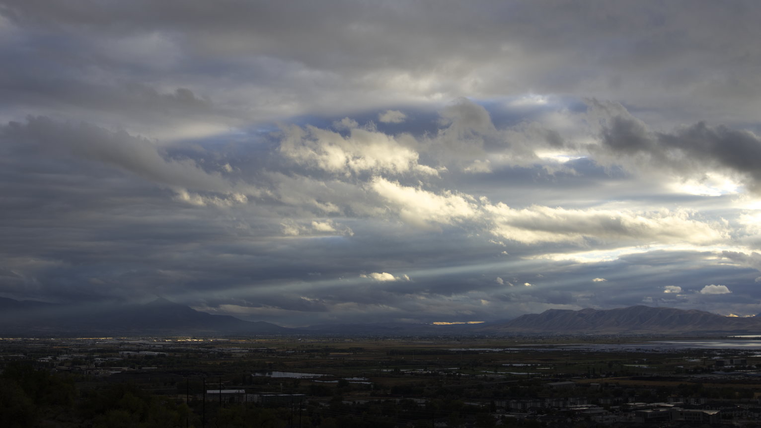 A turquoise ray of sun streaks through the many clouds--some deep grey and blue, others lit brightly by the sun--over town and mountains in the distance