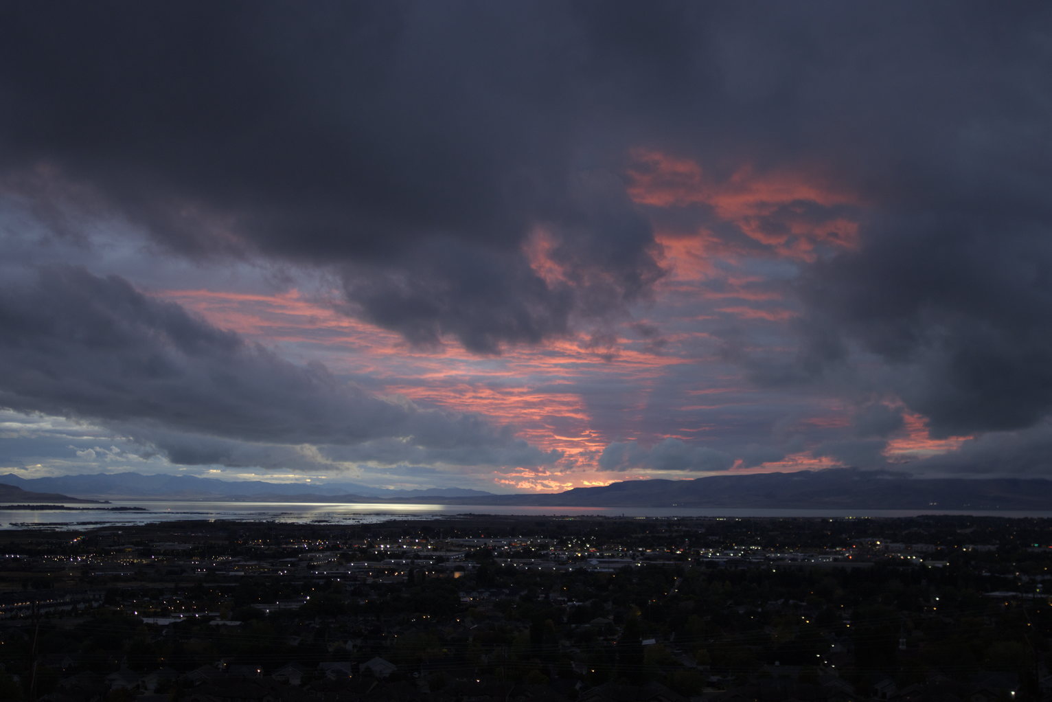 Dramatic red clouds spread across dark blue sky in streaking patterns above the lake and silhouetted mountains, town lights glowing below