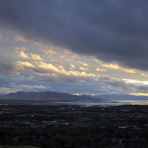 Orange and pink clouds lit by sunset stretch across dark blue sky above Utah Lake and mountains with town lights below