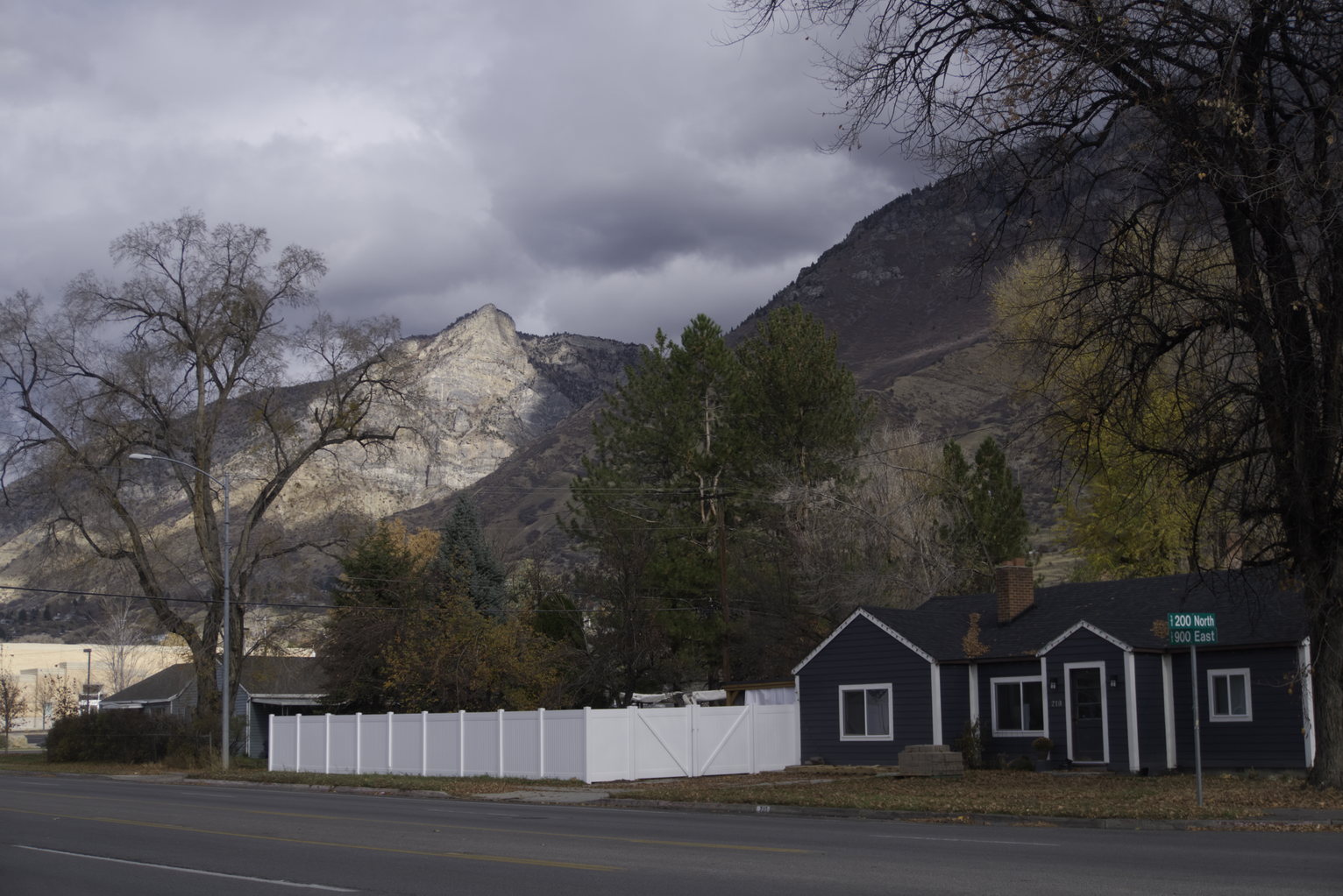 A street corner with a house and white fence, trees, and behind dramatically lit rocky ledge of the mountain, overcast sky