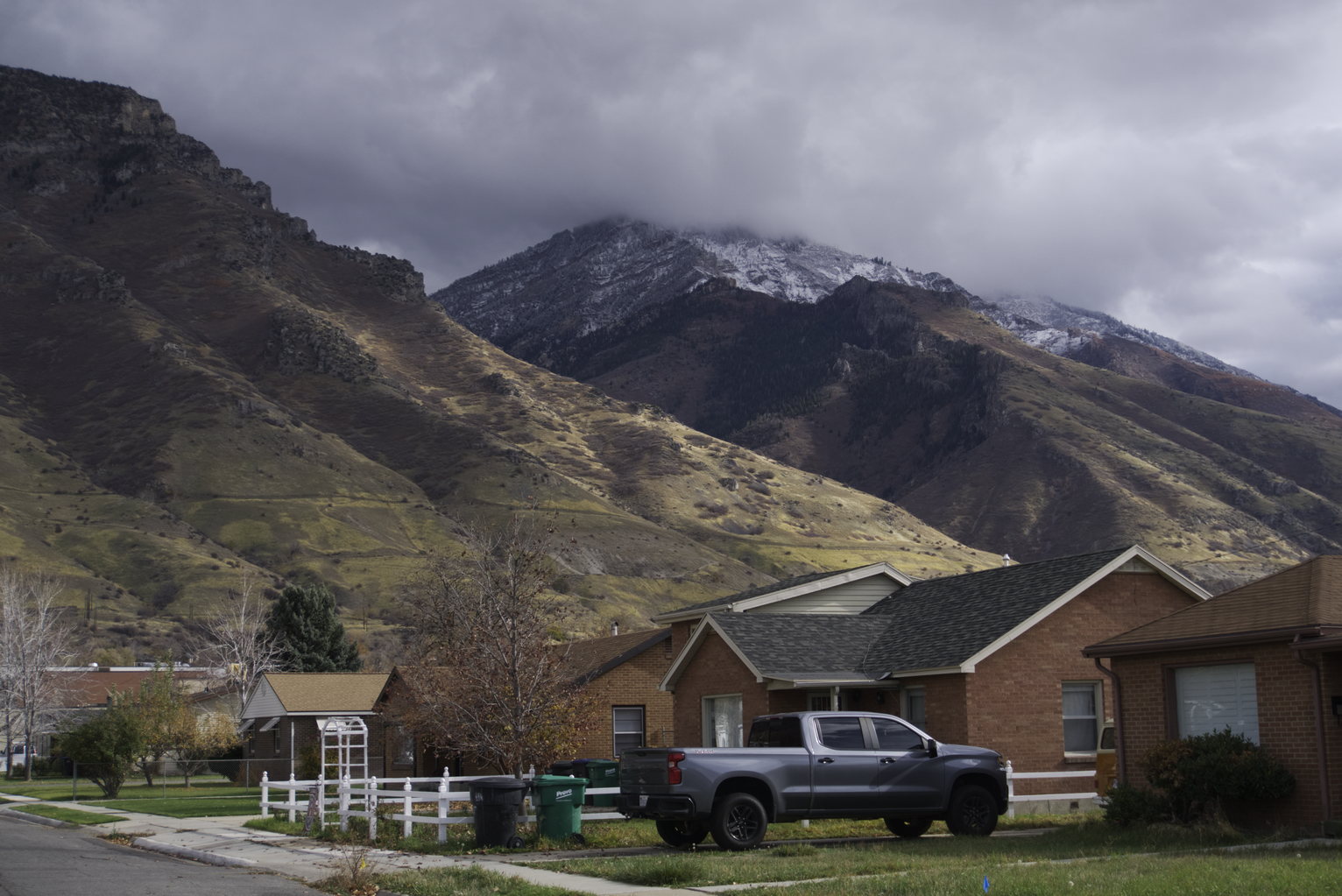 A residential street with houses and trees, the mountainsides softly lit by cloud filtered light, some first powder of snow, overcast skies