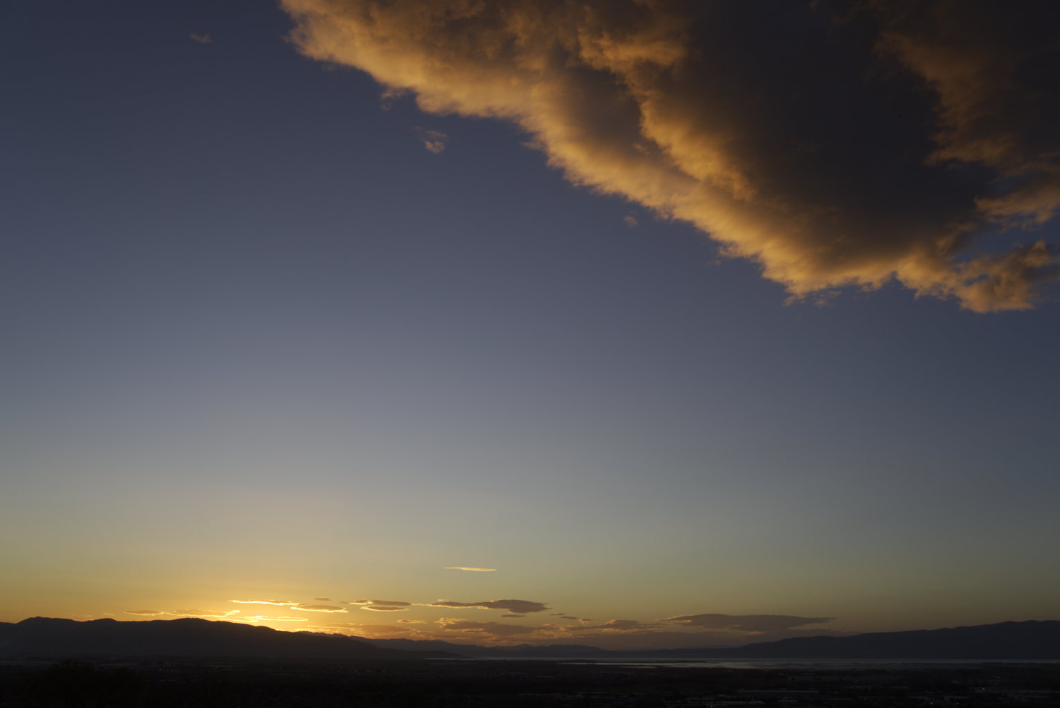 Wide landscape at dusk with golden light glowing on the horizon beneath dark clouds lit orange and gold from below, mountain ridges silhouetted against the fading sky