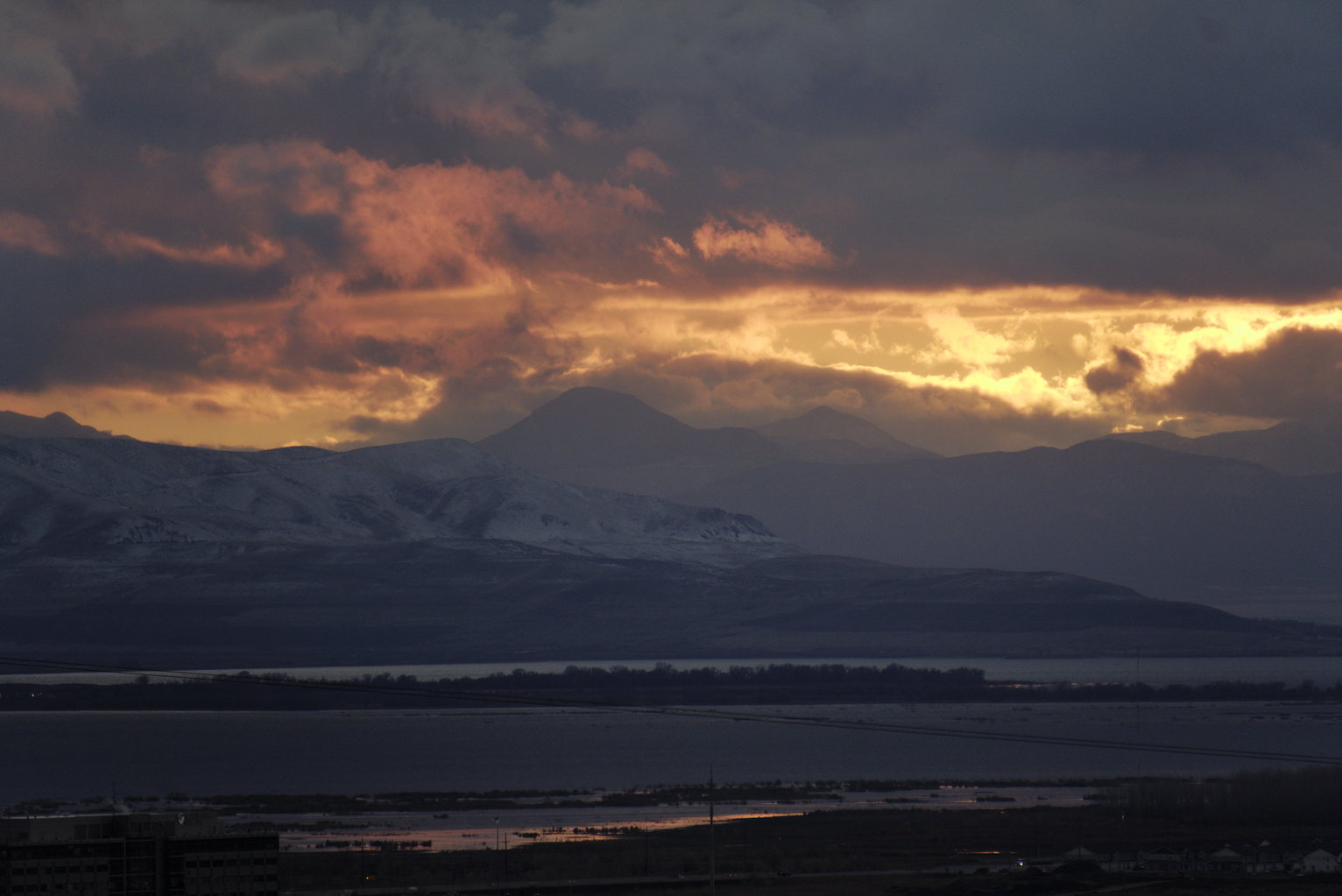 Fiery orange and red clouds illuminated at sunset above dark silhouetted mountain ranges, with a valley and lake below in twilight