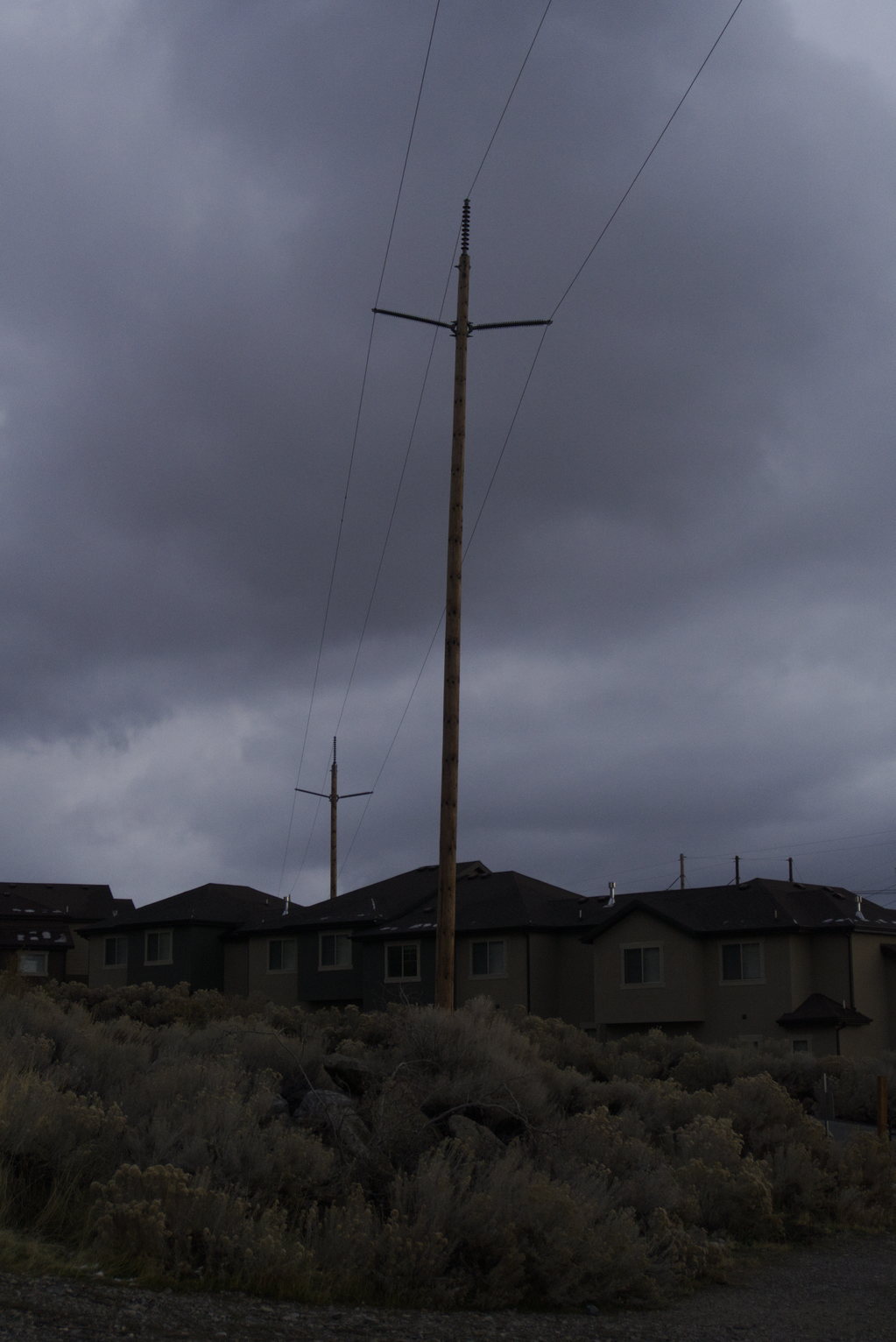 Wooden utility pole with power lines against an overcast twilight sky, with houses and dry brush in the foreground