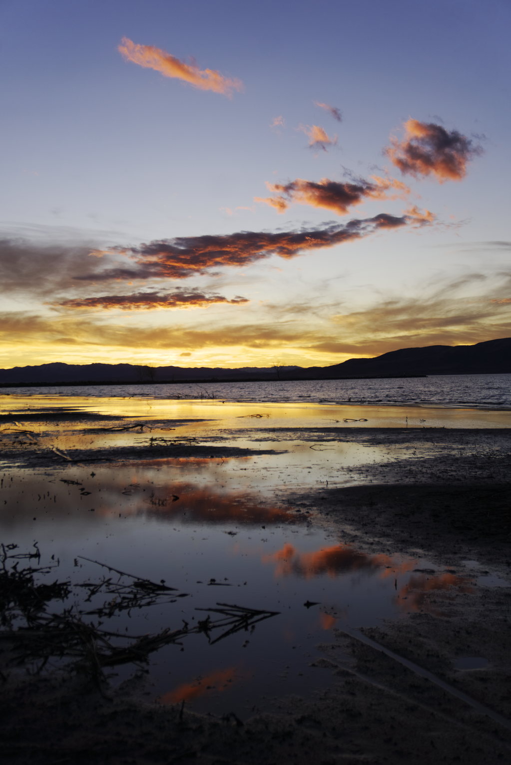 Orange and pink clouds reflected in shallow water and mudflats at sunset, with silhouetted mountains in the distance