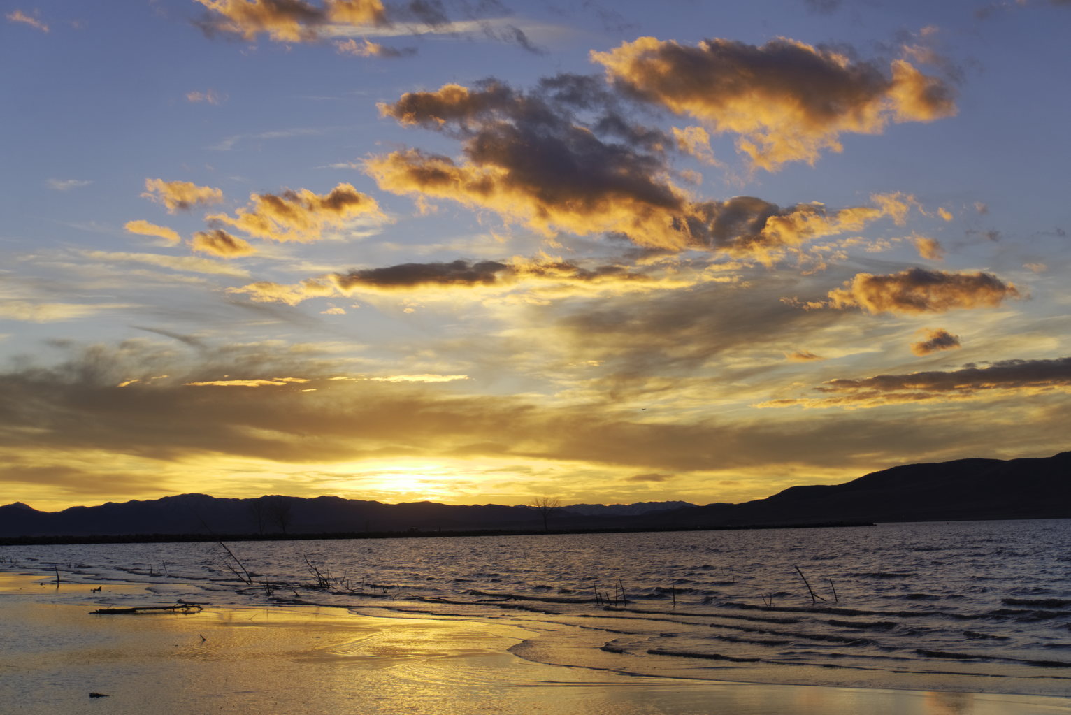 Golden sunset clouds above a lake with dark mountain silhouettes, warm light reflecting on the water's edge