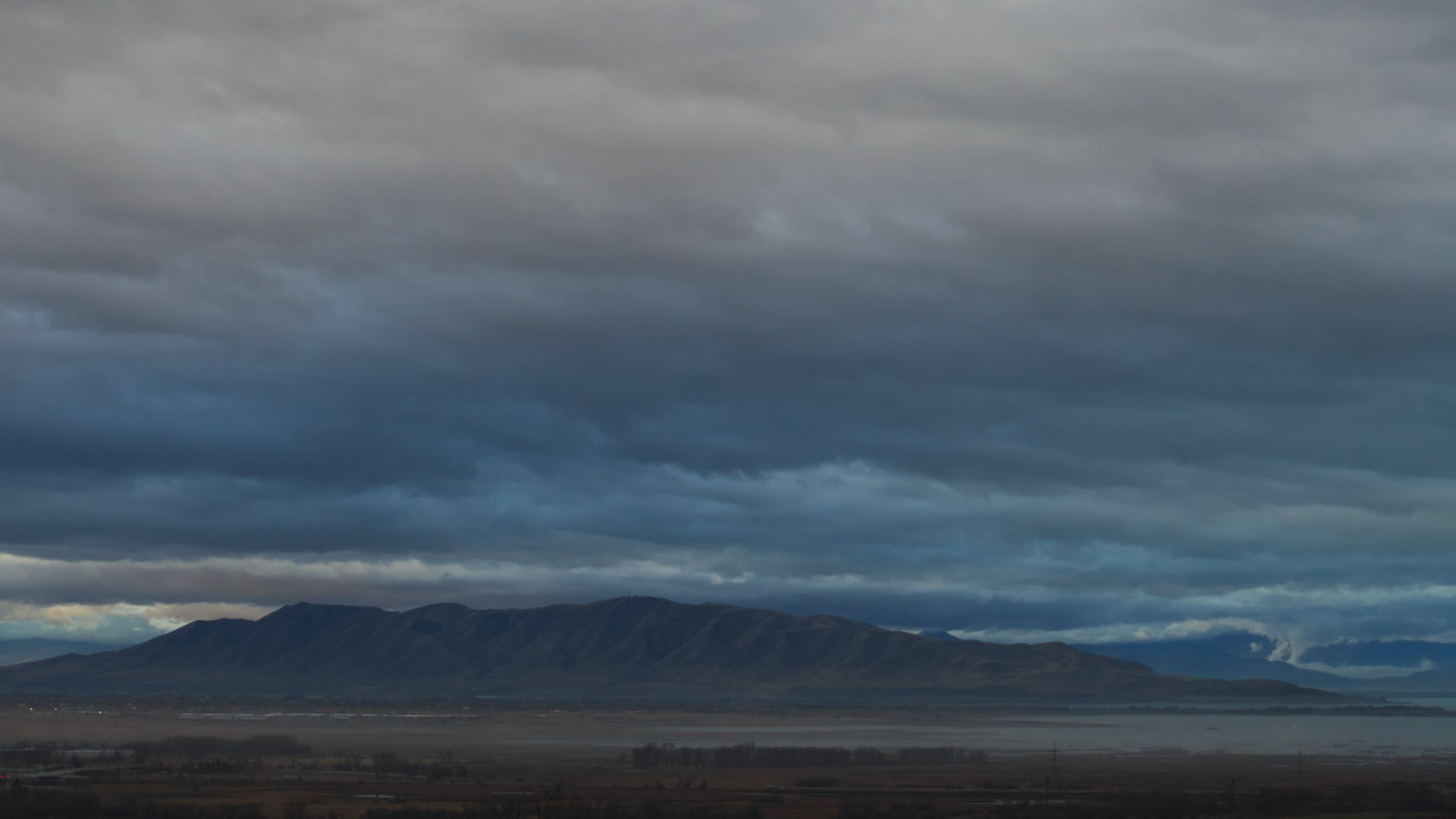 Dramatic gray clouds stretch across a dusky sky above a distant mountain range silhouette, with a valley and scattered lights below