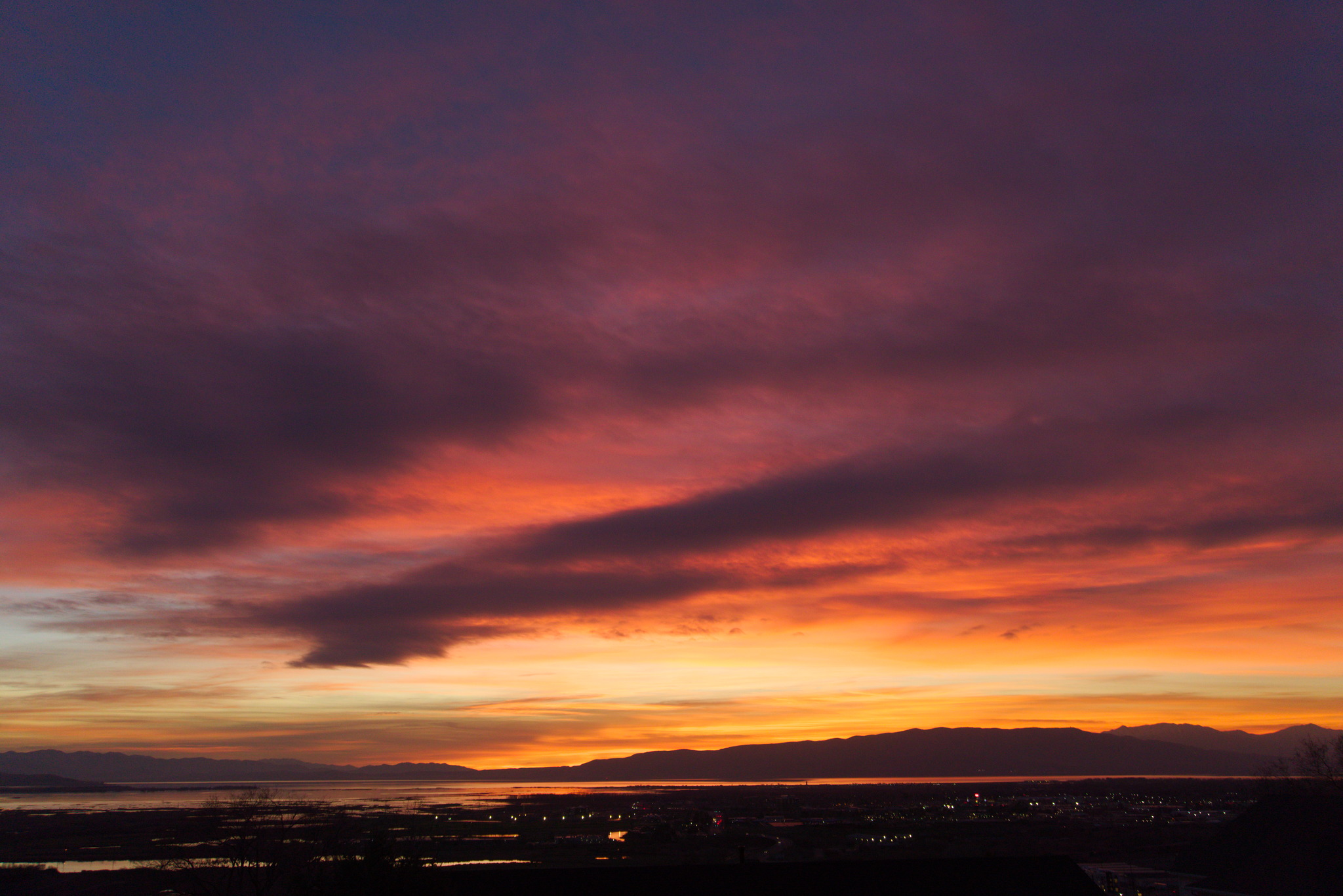 In late sunset, the closer by clouds have turned dark purple grey and the higher ones behind now catch the brilliant red all over a distant mountainline, lake, and town lights