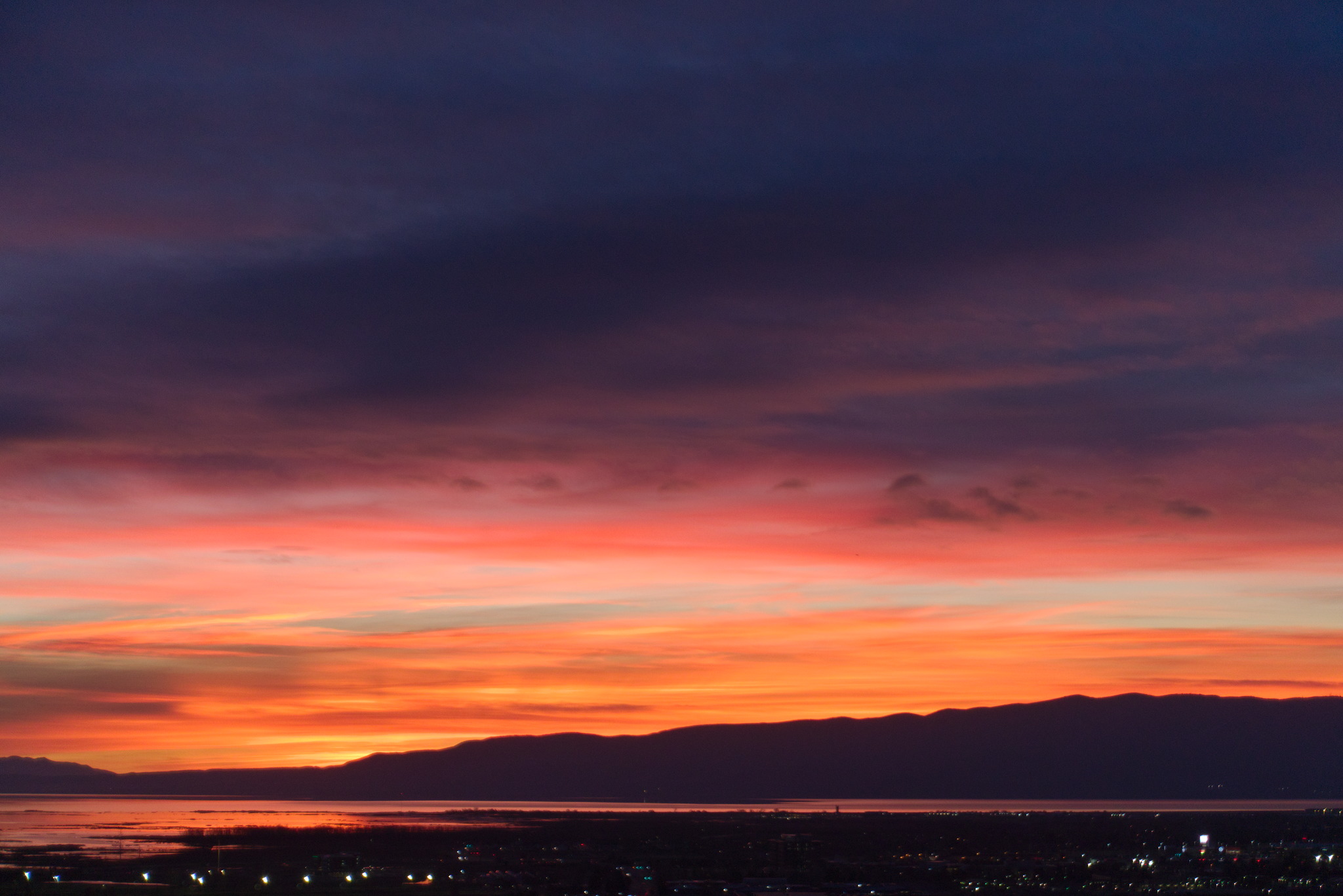 Sunset colors recede and near the horizon the bands of clouds are bright orange, then pink, and then deep blue and purple grey clouds over the distant mountainline, lake, and town lights