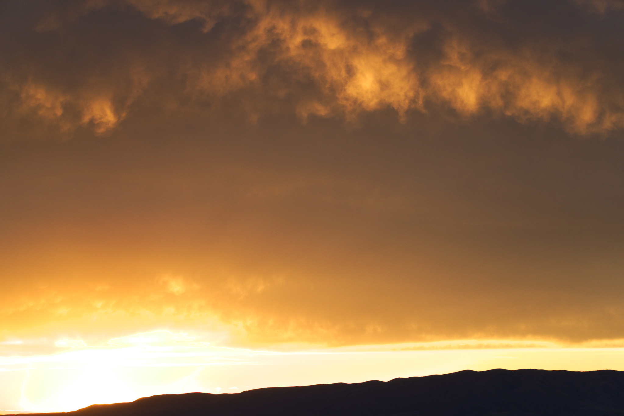 The sun overexposed just above a silhouetted mountainline with clouds catching the light above
