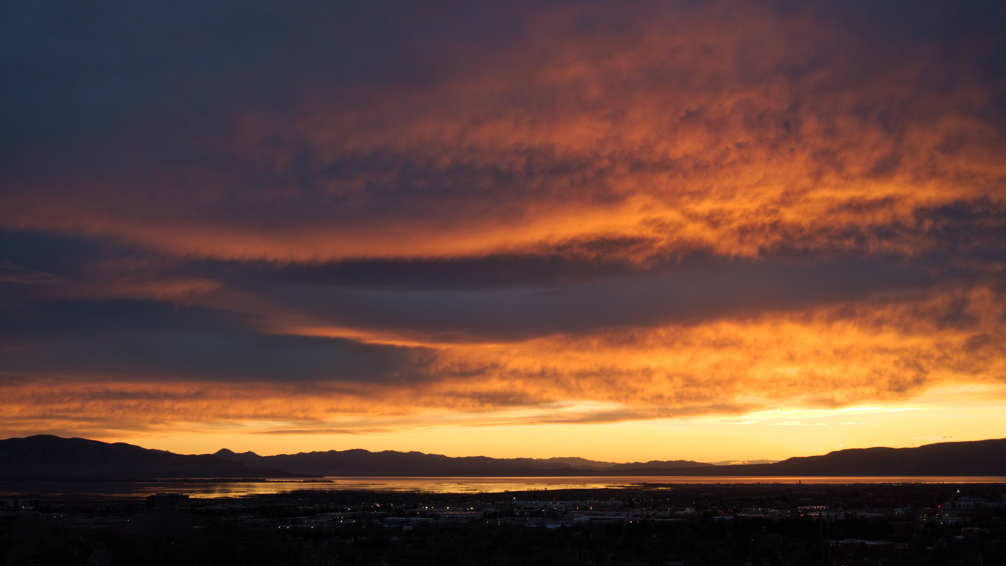 Clouds catching orange light against a backdrop of dark blue-purple clouds above the yellow sky above the distant mountain silhouettes above the lake and town