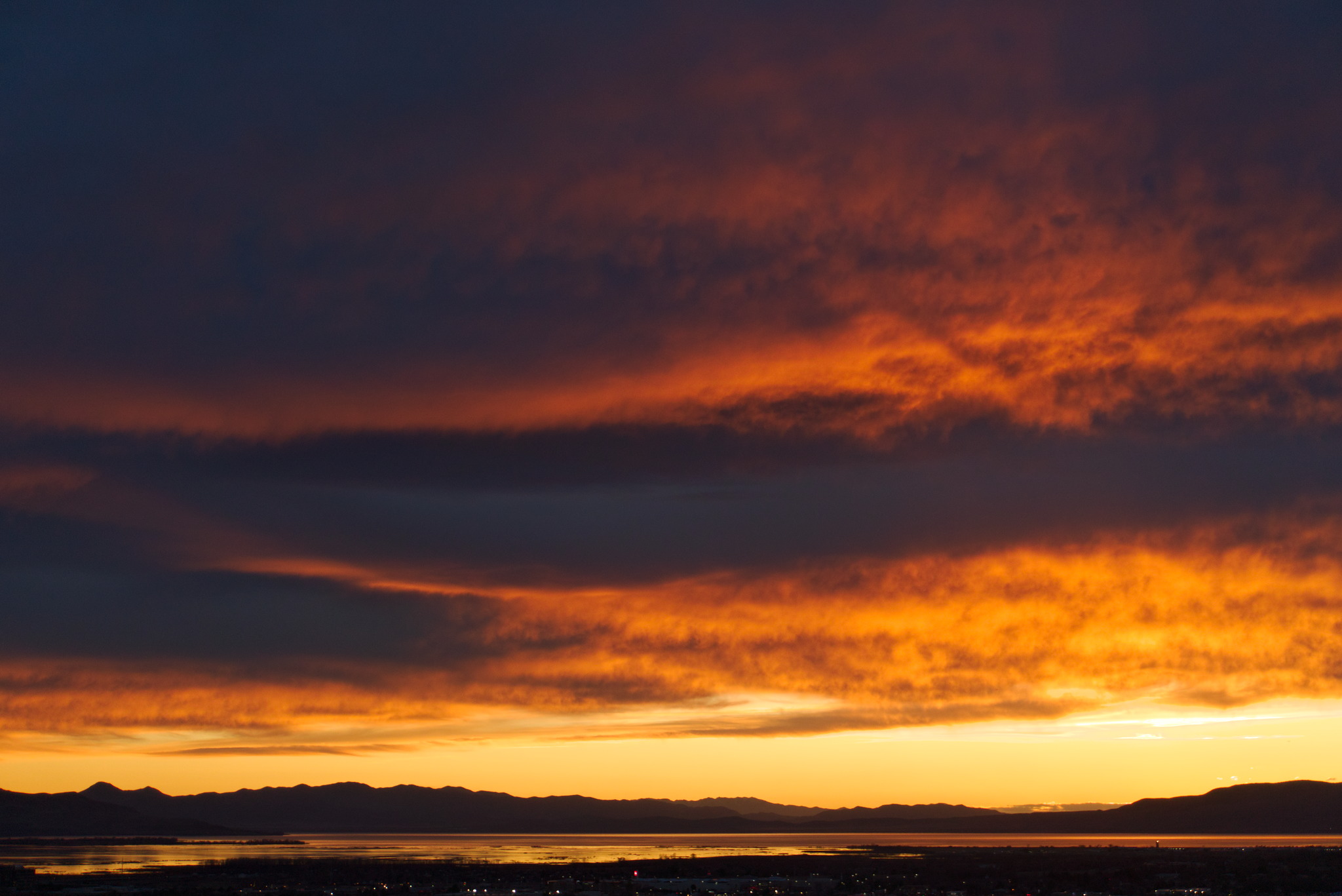 Clouds catch deep orange light against deep dark blue purple clouds in fading sunset above a yellow band of sky just above the distance sillouetted mountains above the lake reflecting orange