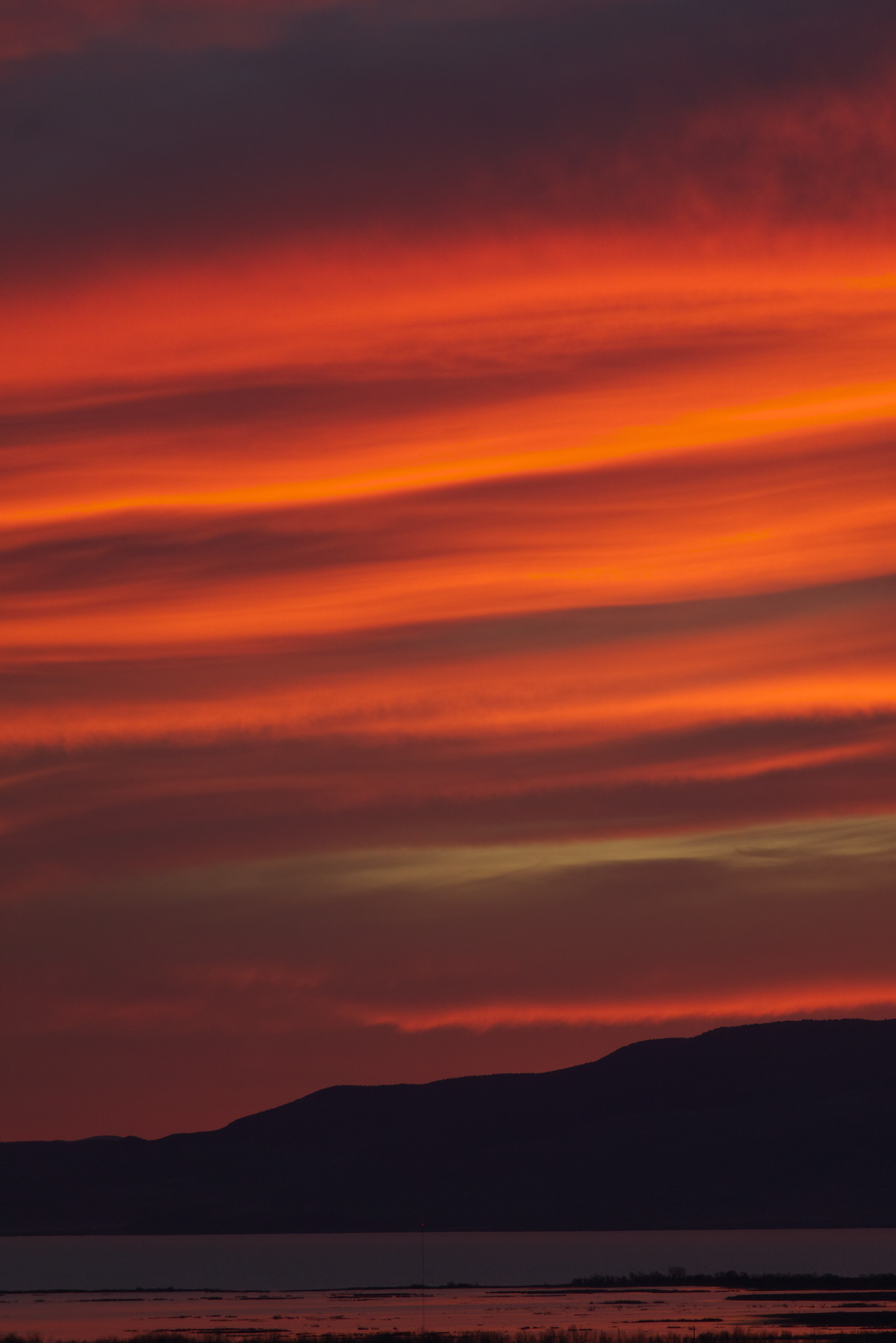 Strongly banded orange sunset lit clouds above silhouetted mountain line above a lake with a reflected band of orange in it