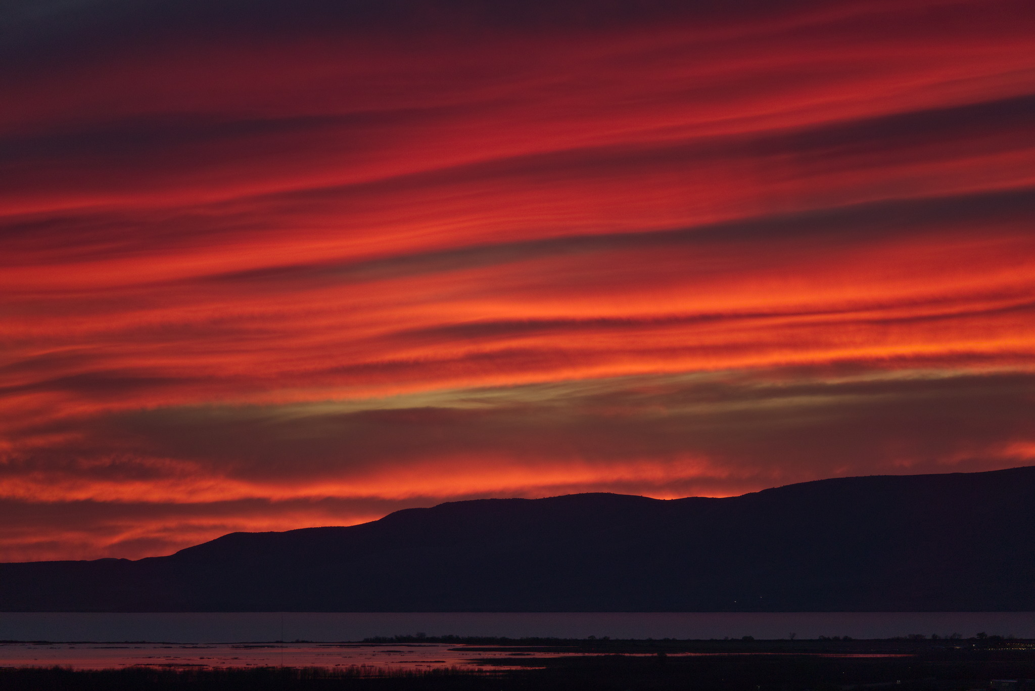Strongly banded orange sunset lit clouds above a silhouetted mountain line above a lake with a reflected orange band
