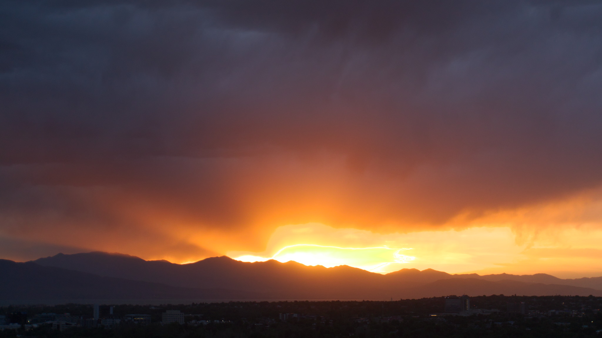 Orange sunset streams up into dark grey clouds over a distant mountainline and a dark town
