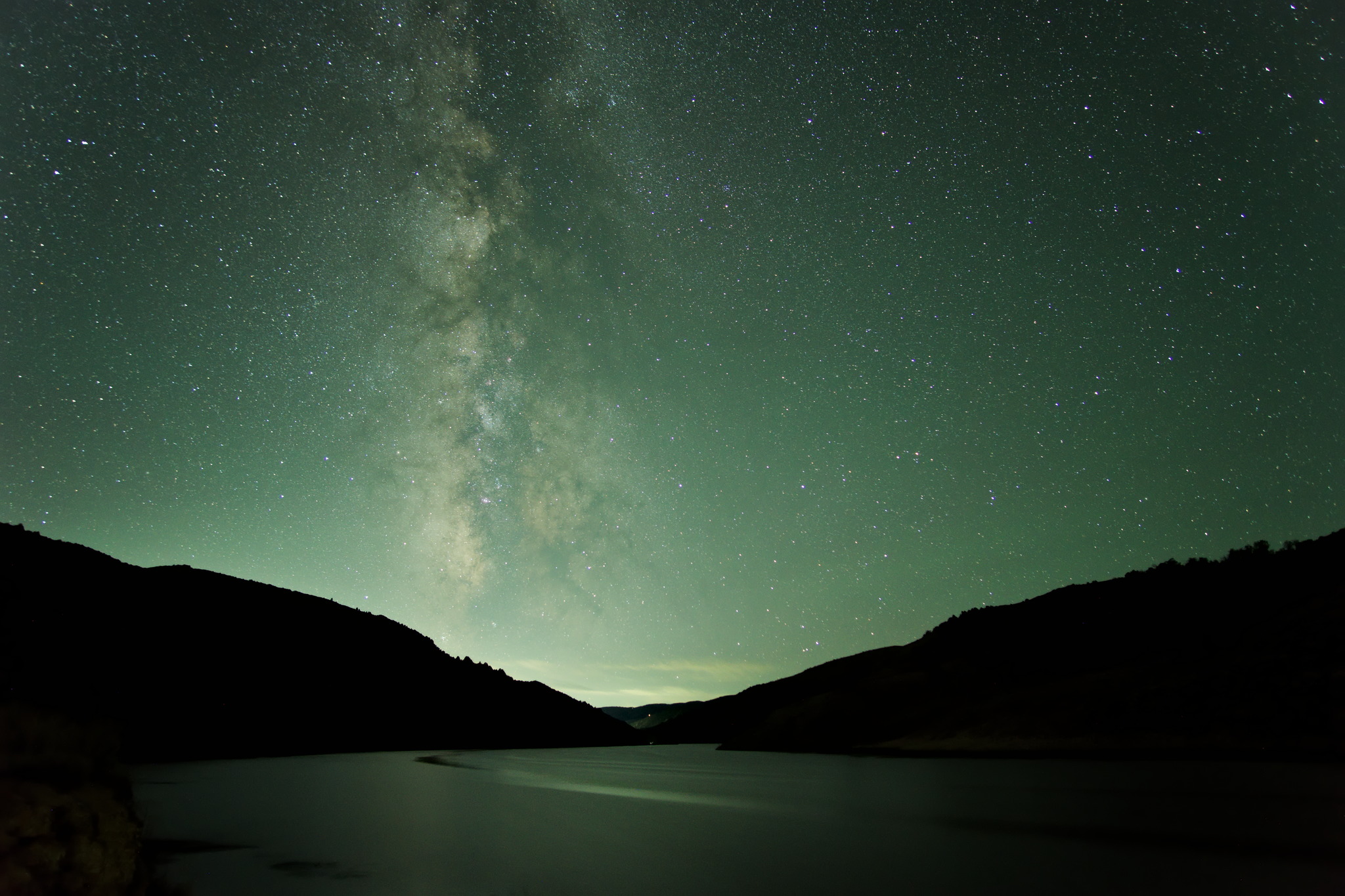 A nightscape featuring the milky way rising vertically in greenish starry night sky above a river gleaming with the same greenish light