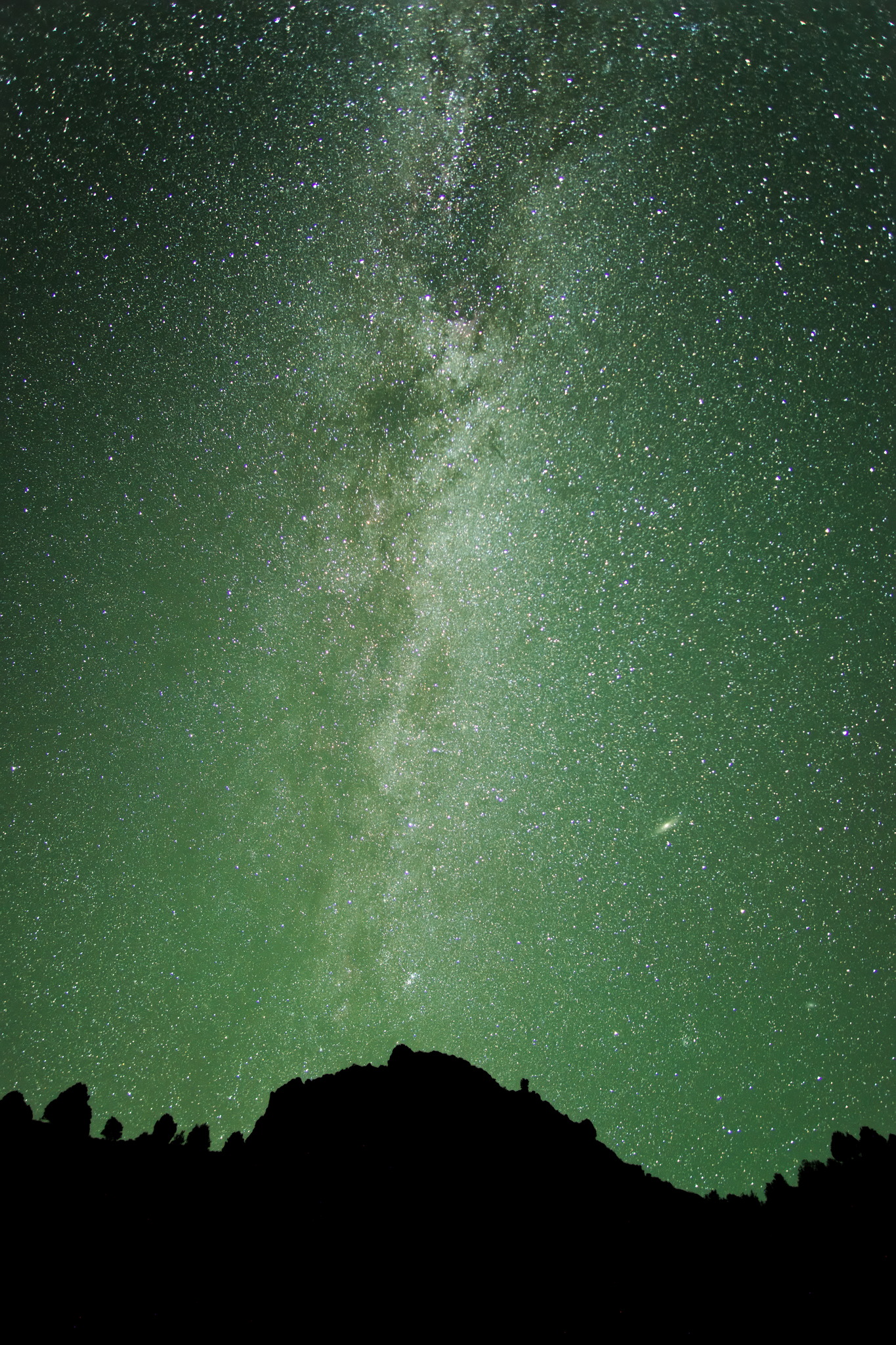 The milky way rises bright out of the silhouette of a nearby hill, the sky is green with airglow