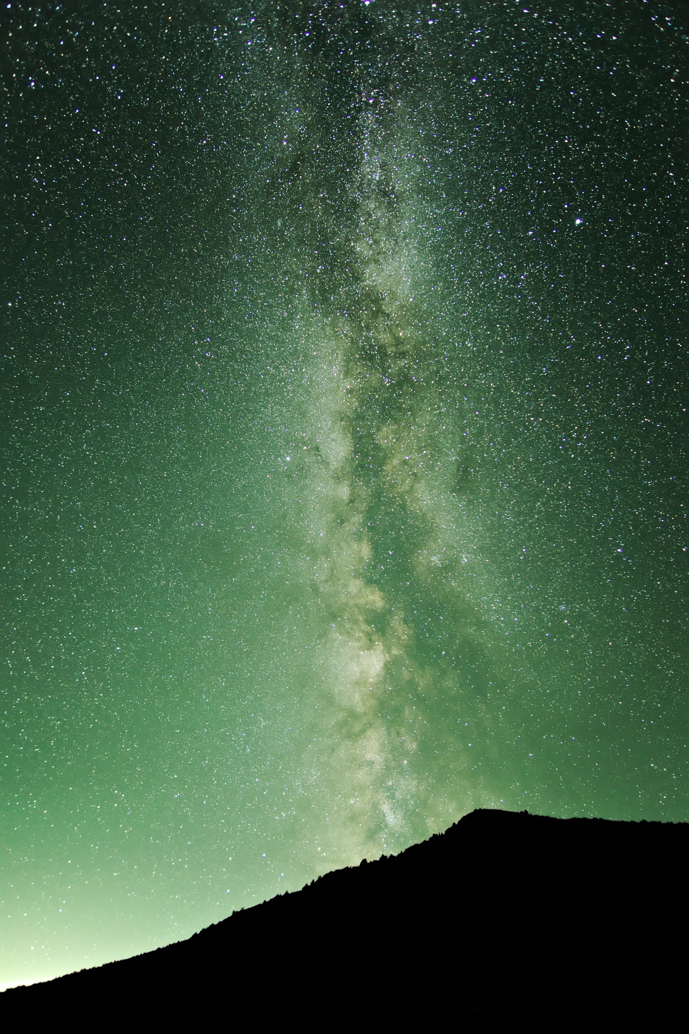 The milky way rises bright out of an angular silhouetted hill, the sky nightsky glows green with airglow