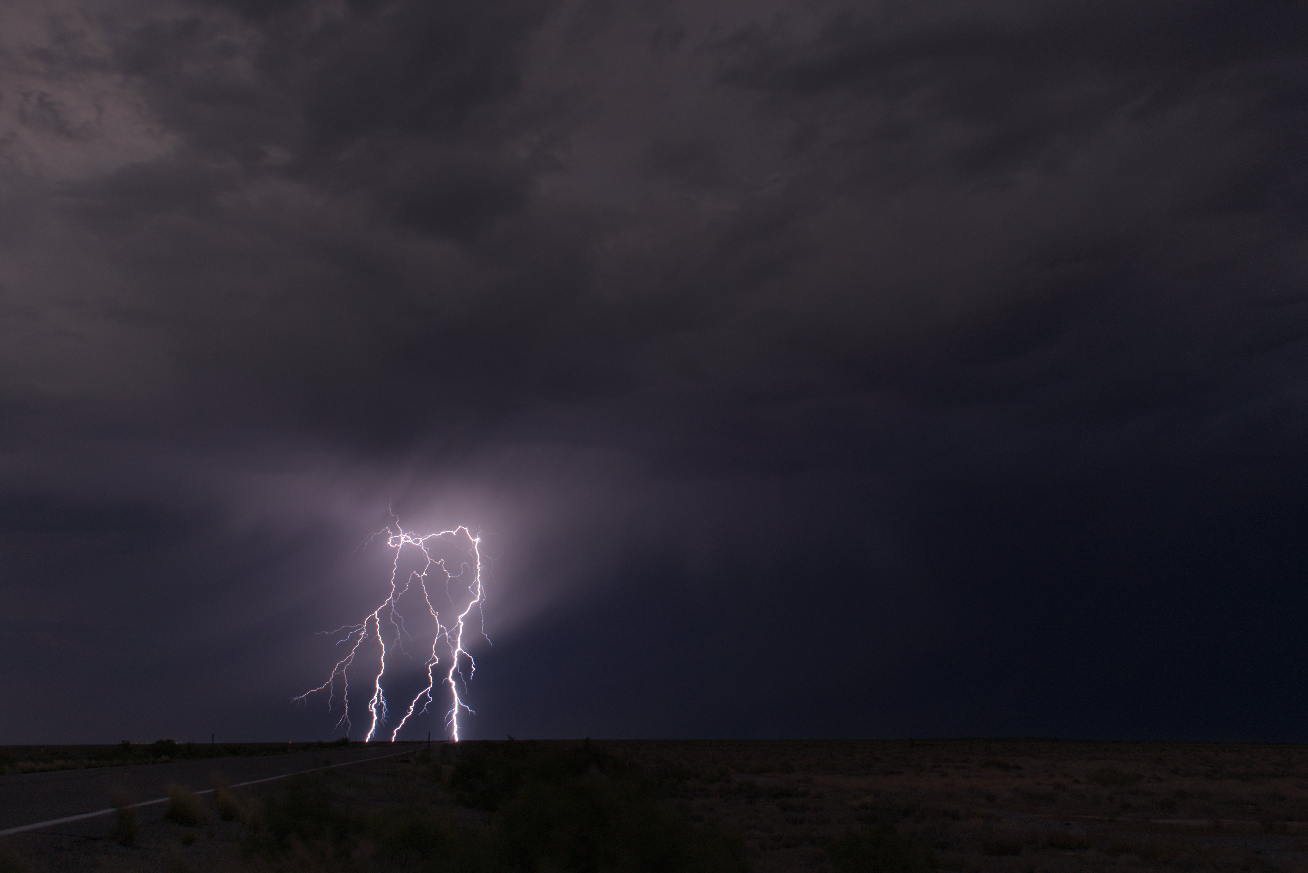 Dramatic lightning bolts illuminate dark storm clouds over a rural landscape with a road in the foreground and distant mountains silhouetted against the stormy sky.