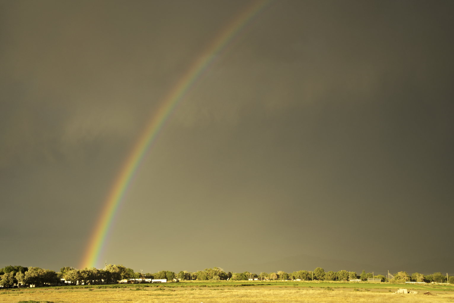 A vibrant rainbow arcs across golden fields and rural landscape with scattered buildings and trees under dramatic cloudy skies.