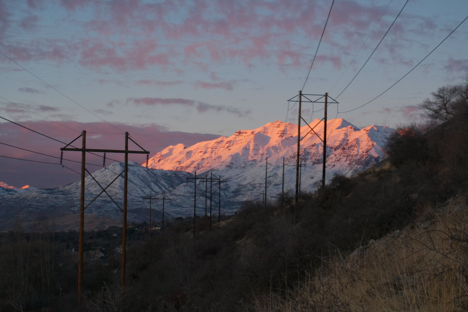 The upper half of a mountain bathed in sunset light, pinky clouds above, and in the foreground powerlines running forward.