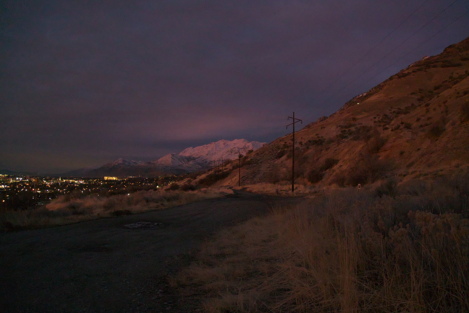 The mountainside seems to glow a warm light in the late dusk, snowy mountains in the distance, some city lights.