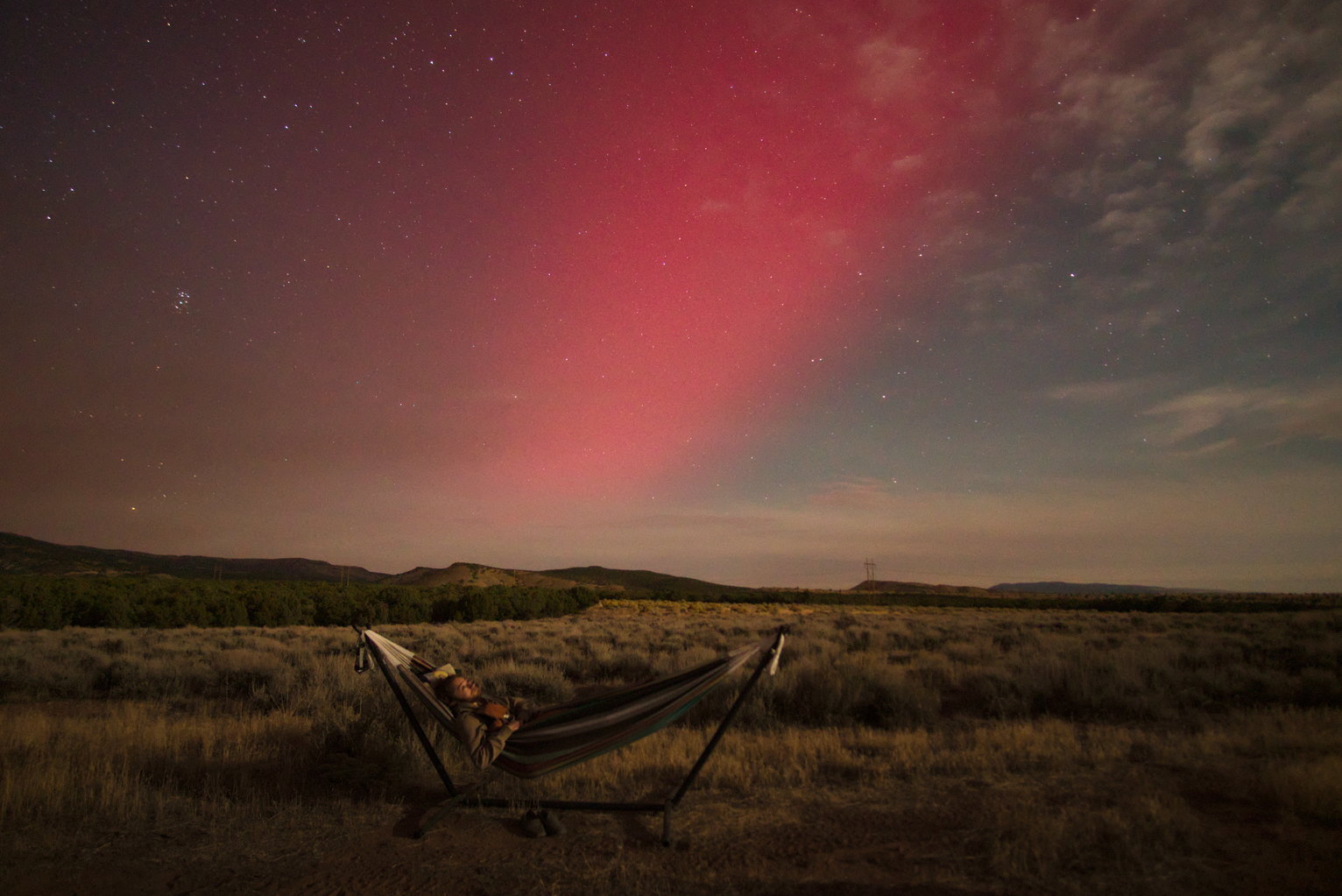 Someone lays in a hammock looking at the night sky which has a pink aurora swash in it